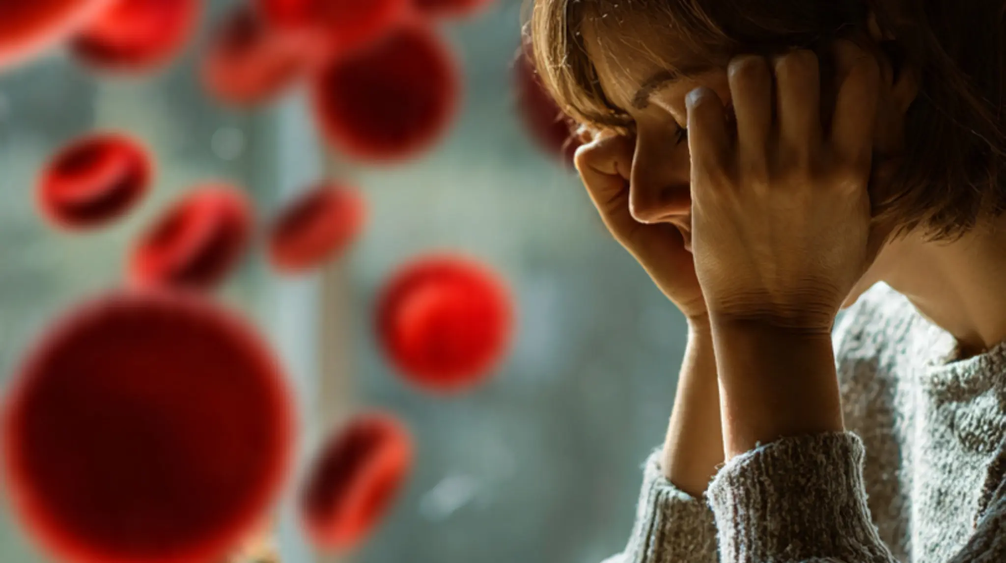 A fatigued woman leaning her head on her hands with floating red blood cell graphics, illustrating the symptoms of iron deficiency anemia.