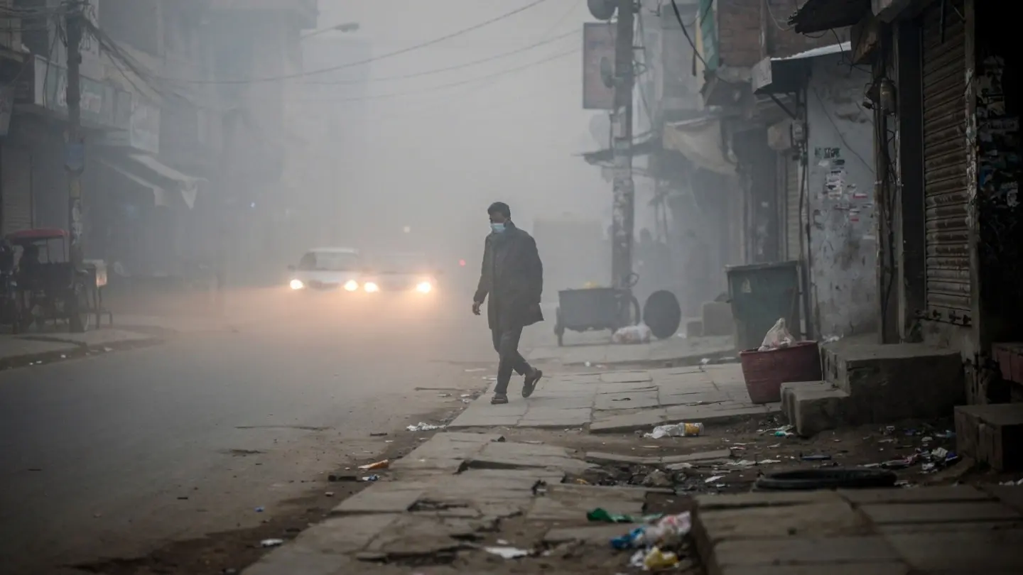 A man wearing a face mask walks down a heavily smoggy street in Pakistan, illustrating the health risks of seasonal illness temperature shifts.